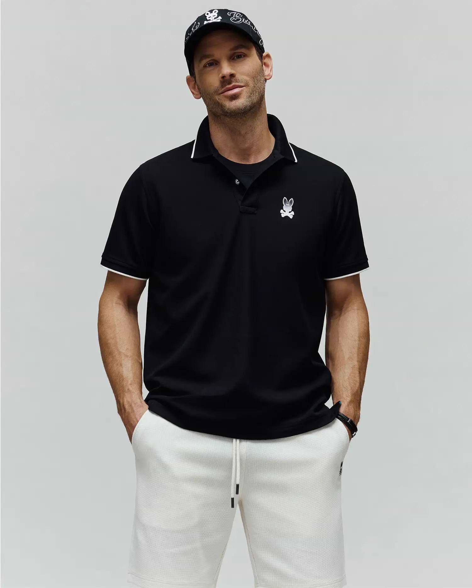 Mens Aram Pique Polo, a black classic fit pique knit polo shirt with white contrast tipping on the collar and sleeve cuffs, featuring an embroidered white Psycho Bunny skull logo on the left chest. (1)