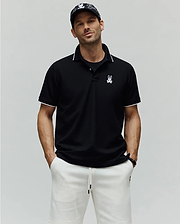 Mens Aram Pique Polo, a black classic fit pique knit polo shirt with white contrast tipping on the collar and sleeve cuffs, featuring an embroidered white Psycho Bunny skull logo on the left chest. (1)