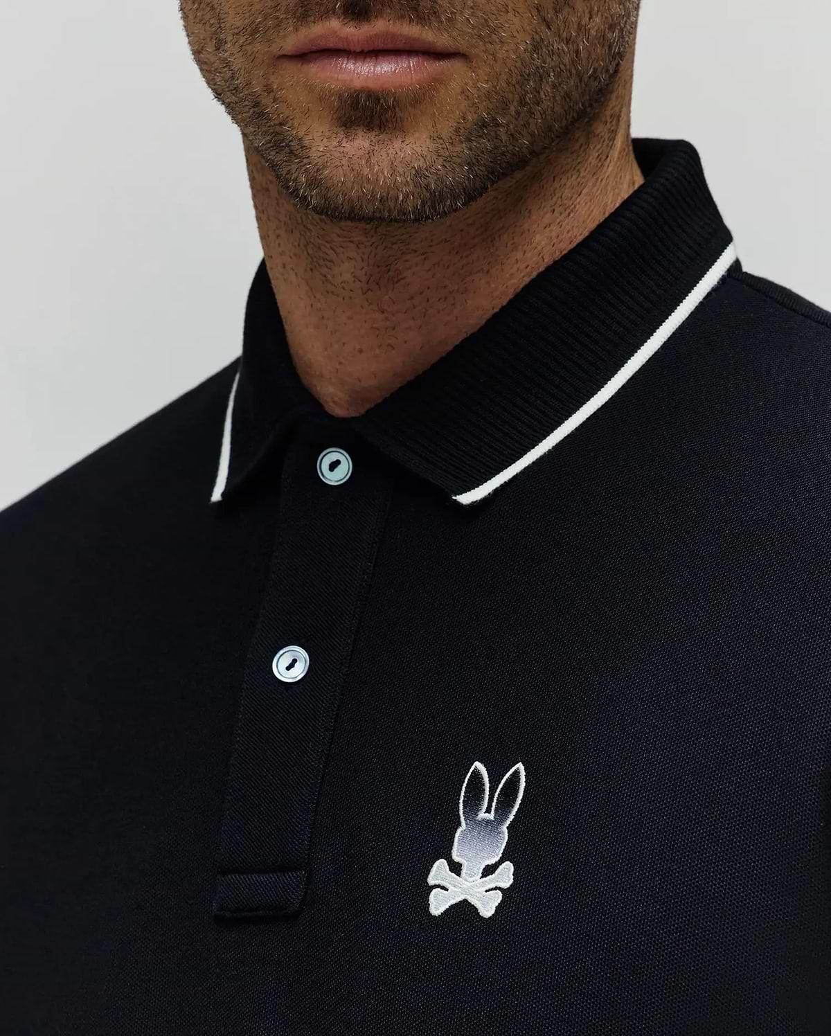 Black Mens Aram Pique Polo with a classic fit, featuring a white-tipped ribbed collar, two light blue buttons, and a medium embroidered white and grey Psycho Bunny skull logo on the left chest. (2)