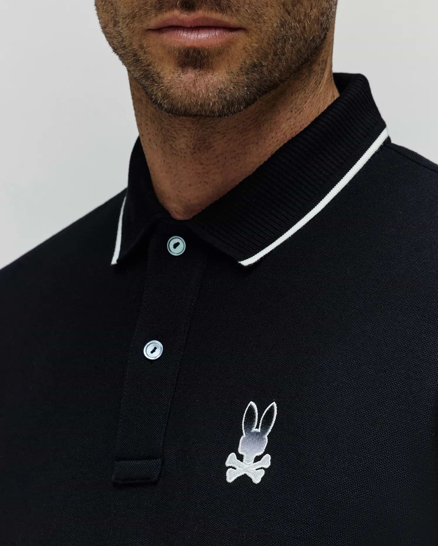 Black Mens Aram Pique Polo with a classic fit, featuring a white-tipped ribbed collar, two light blue buttons, and a medium embroidered white and grey Psycho Bunny skull logo on the left chest. (2)