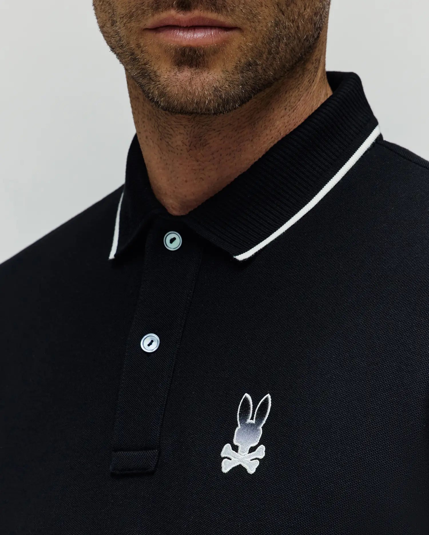 Black Mens Aram Pique Polo with a classic fit, featuring a white-tipped ribbed collar, two light blue buttons, and a medium embroidered white and grey Psycho Bunny skull logo on the left chest. (2)