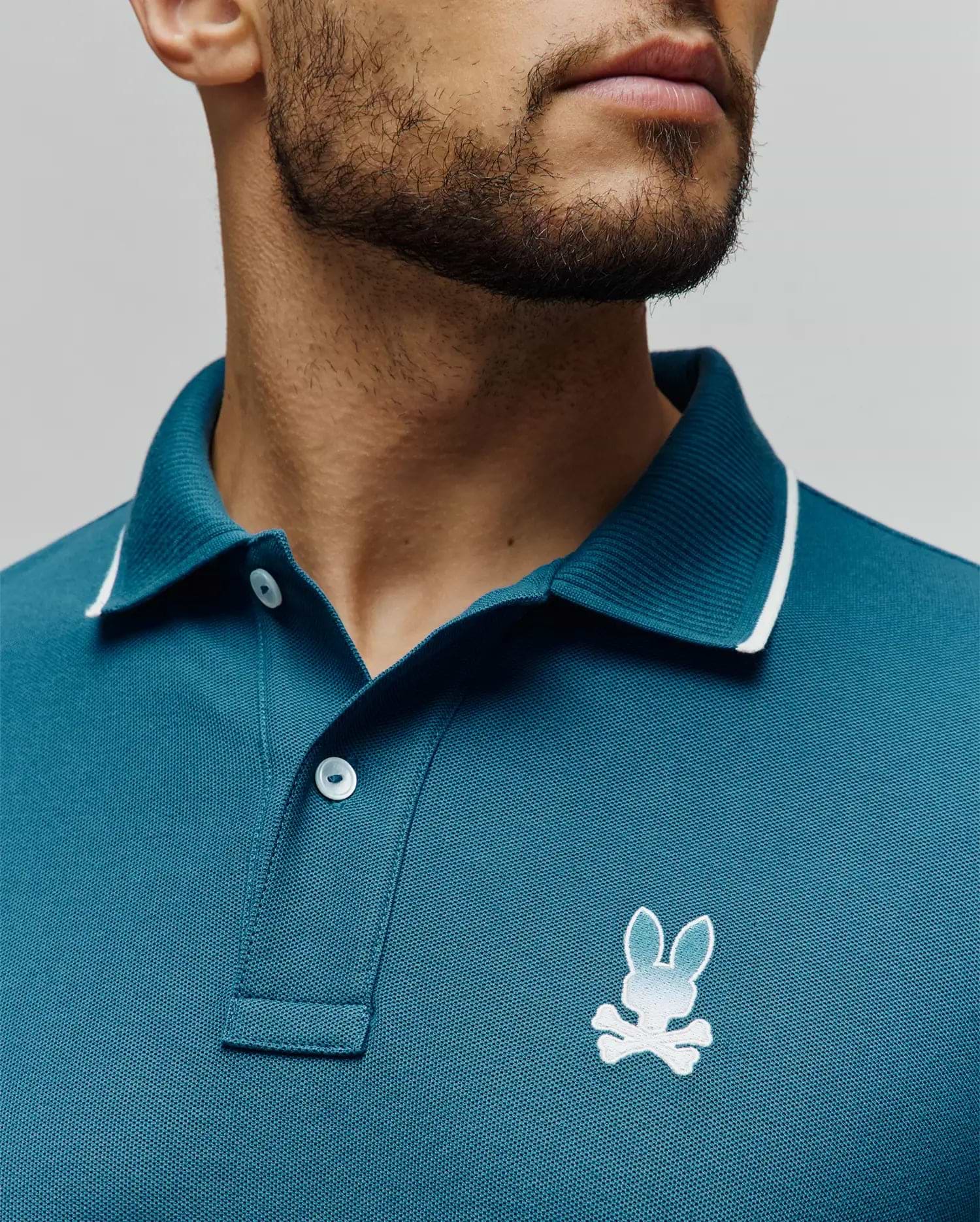 Mens Aram Pique Polo in teal, classic fit, pique knit, with a ribbed collar featuring white tipping, two white buttons, and a small embroidered white to light blue gradient Psycho Bunny skull logo on the left chest. (3)