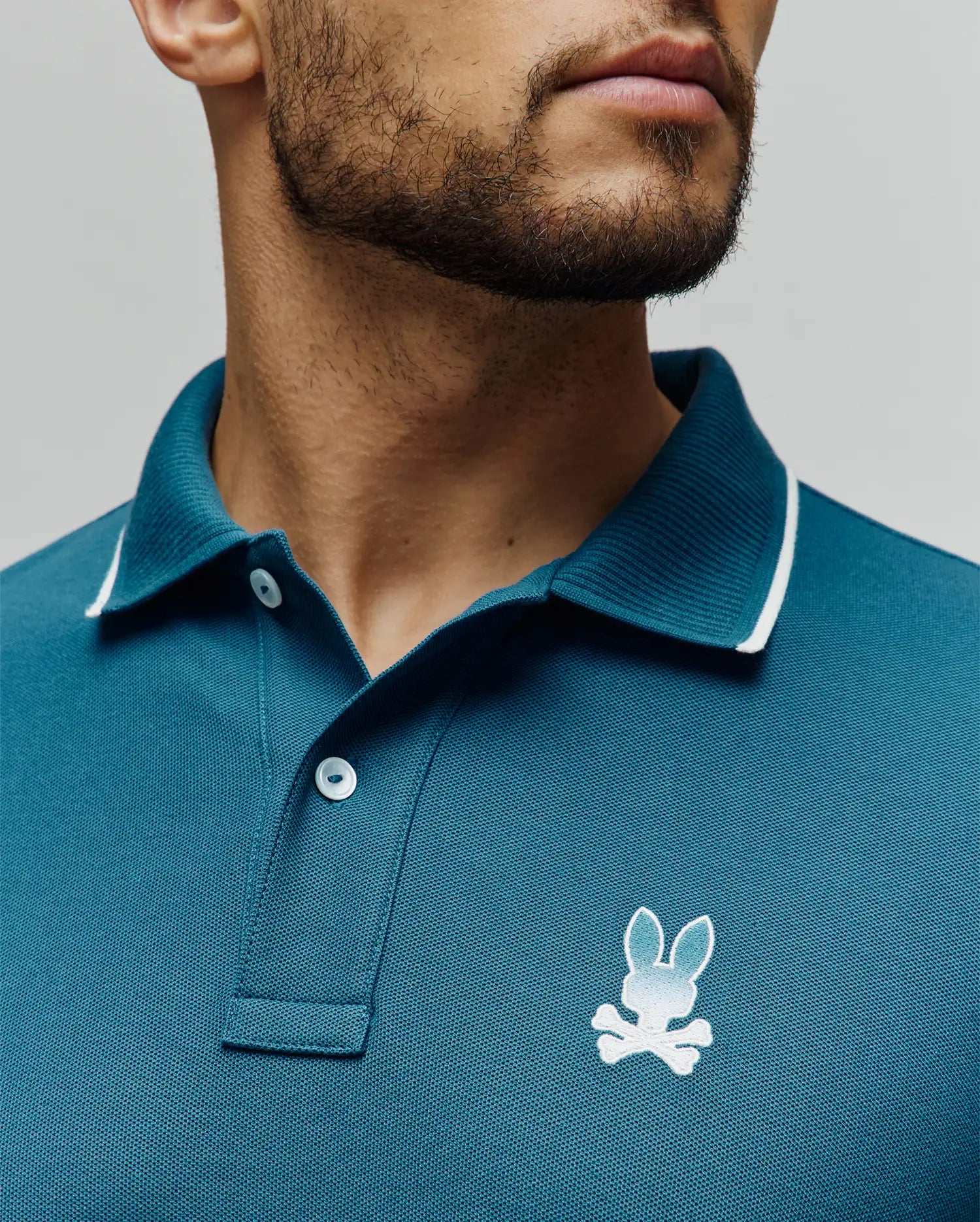 Mens Aram Pique Polo in teal, classic fit, pique knit, with a ribbed collar featuring white tipping, two white buttons, and a small embroidered white to light blue gradient Psycho Bunny skull logo on the left chest. (3)