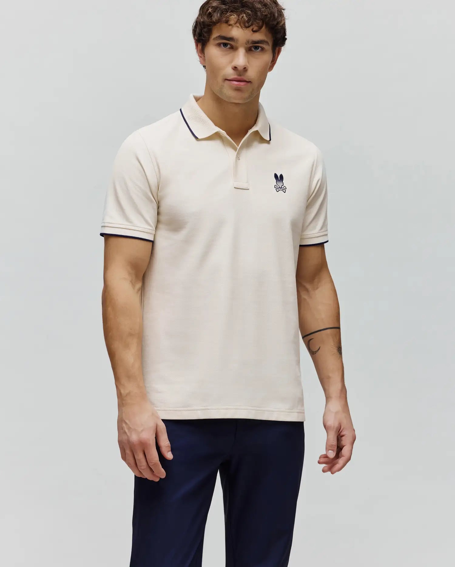 Mens Aram Pique Polo in cream, featuring a classic fit, textured pique fabric, navy blue tipping on the collar and sleeve cuffs, and a small embroidered Psycho Bunny skull logo on the left chest. (3)
