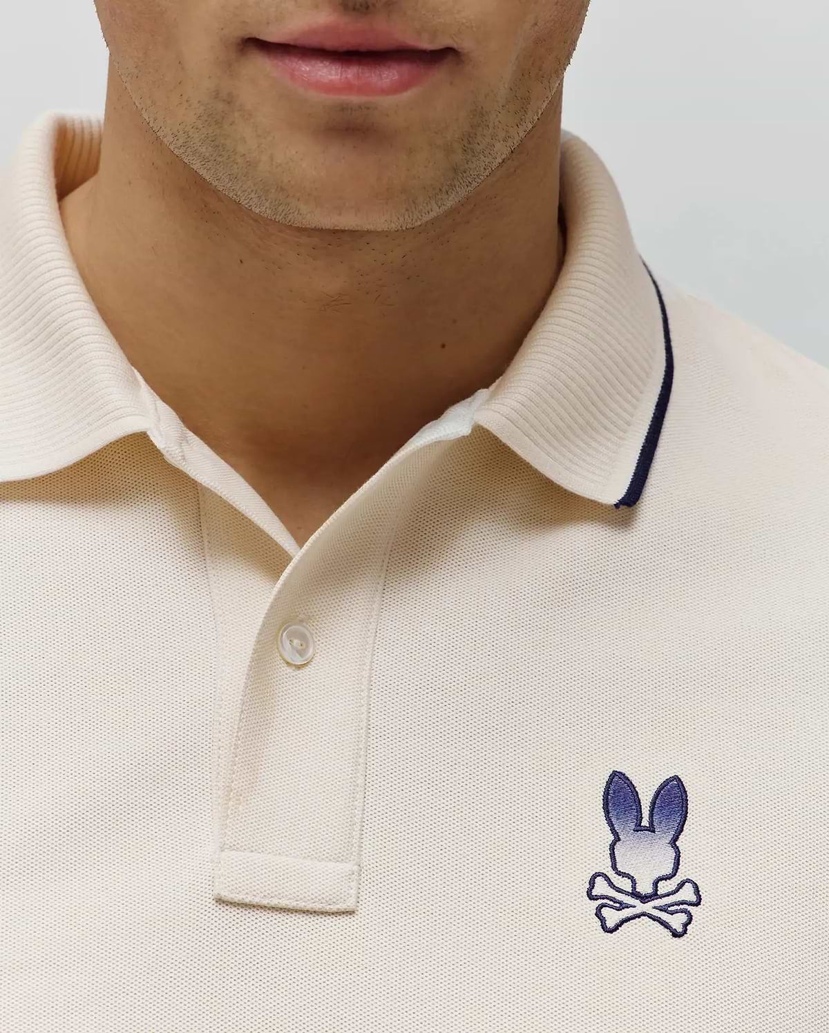 Mens Aram Pique Polo in cream, featuring a pique knit texture, ribbed collar with navy tipping, two-button placket, and an embroidered blue gradient Psycho Bunny skull logo on the left chest. (2)