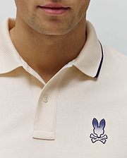 Mens Aram Pique Polo in cream, featuring a pique knit texture, ribbed collar with navy tipping, two-button placket, and an embroidered blue gradient Psycho Bunny skull logo on the left chest. (2)
