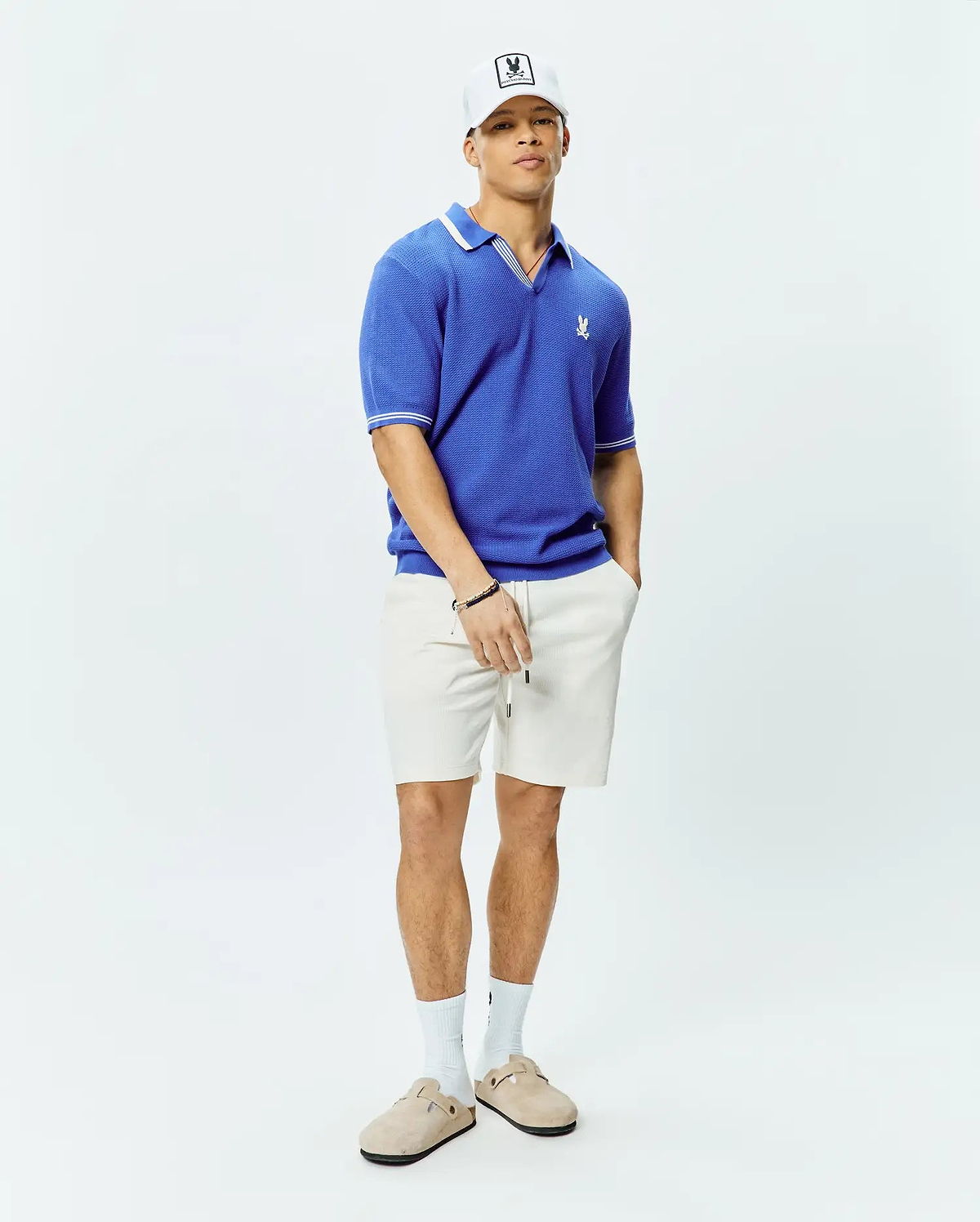 Mens Innis Polo, a vibrant blue classic fit textured knit polo with white tipped collar and cuffs, featuring a small embroidered Psycho Bunny skull logo on the left chest, and relaxed off-white textured drawstring shorts. (4)