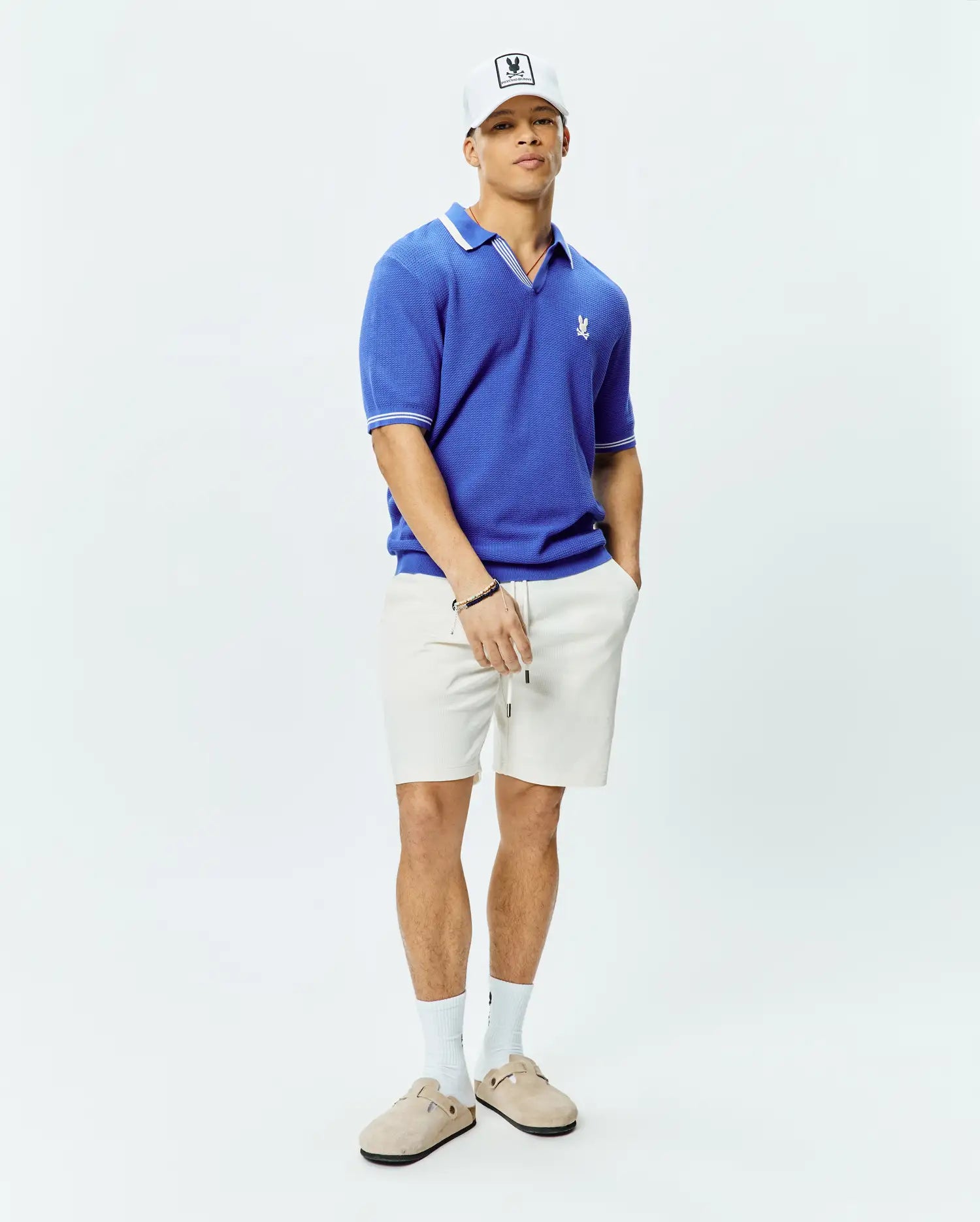 Mens Innis Polo, a vibrant blue classic fit textured knit polo with white tipped collar and cuffs, featuring a small embroidered Psycho Bunny skull logo on the left chest, and relaxed off-white textured drawstring shorts. (4)