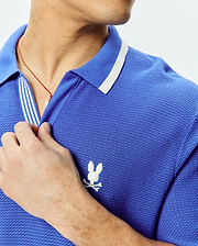 Mens Innis Polo, vibrant blue pique knit polo shirt with a classic fit, featuring a white striped collar, striped placket, and a small white embroidered Psycho Bunny skull logo on the left chest. (2)