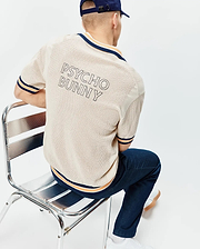 Mens Theros Knit Polo in beige, featuring a breathable open knit texture, classic fit, navy and orange striped ribbed collar, cuffs, and hem, with a large PSYCHO BUNNY text logo on the back. (1)