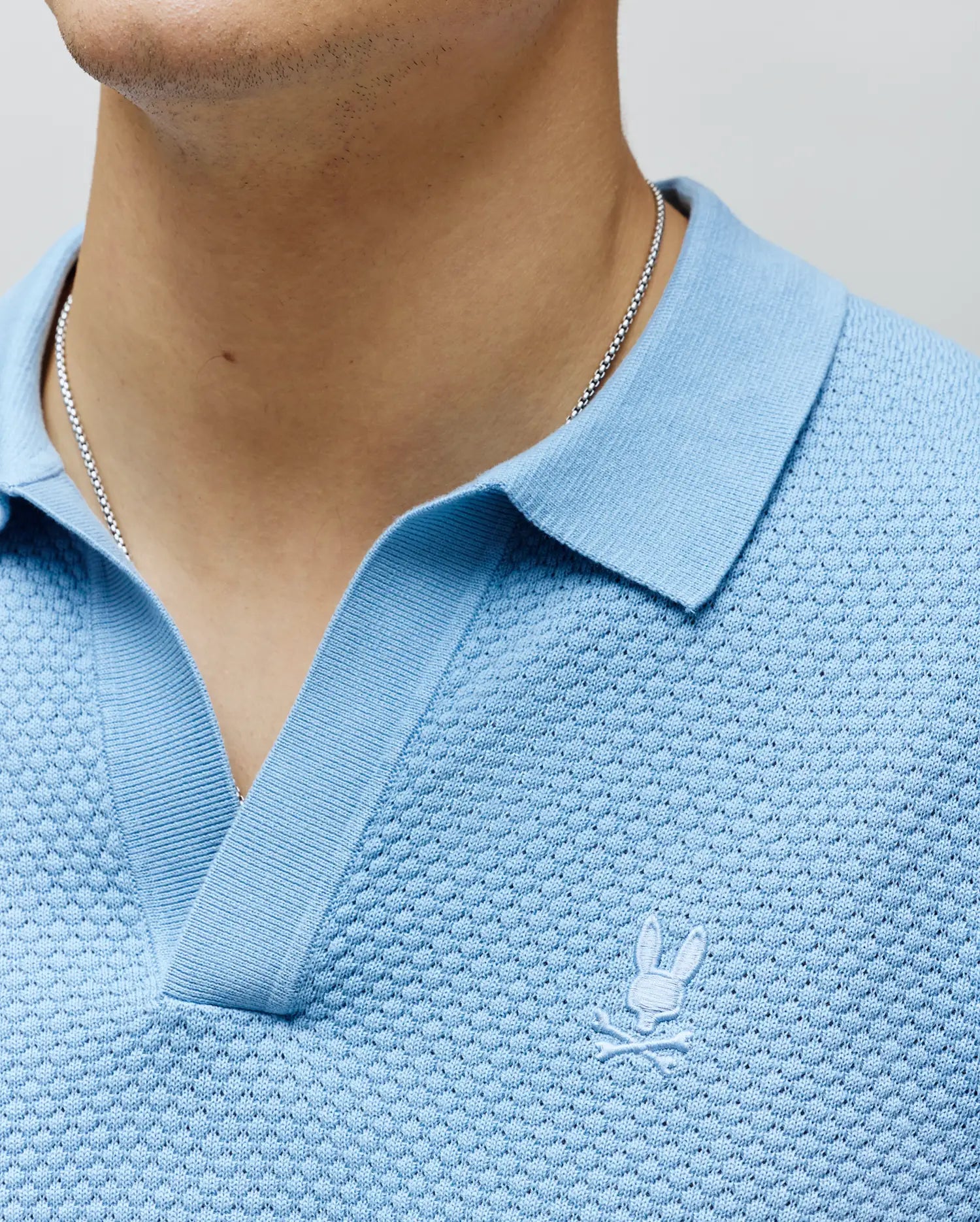 Mens Devon Knit Polo in light blue, featuring a textured knit fabric, ribbed collar, V-neck placket, and a small white embroidered Psycho Bunny skull logo on the left chest.