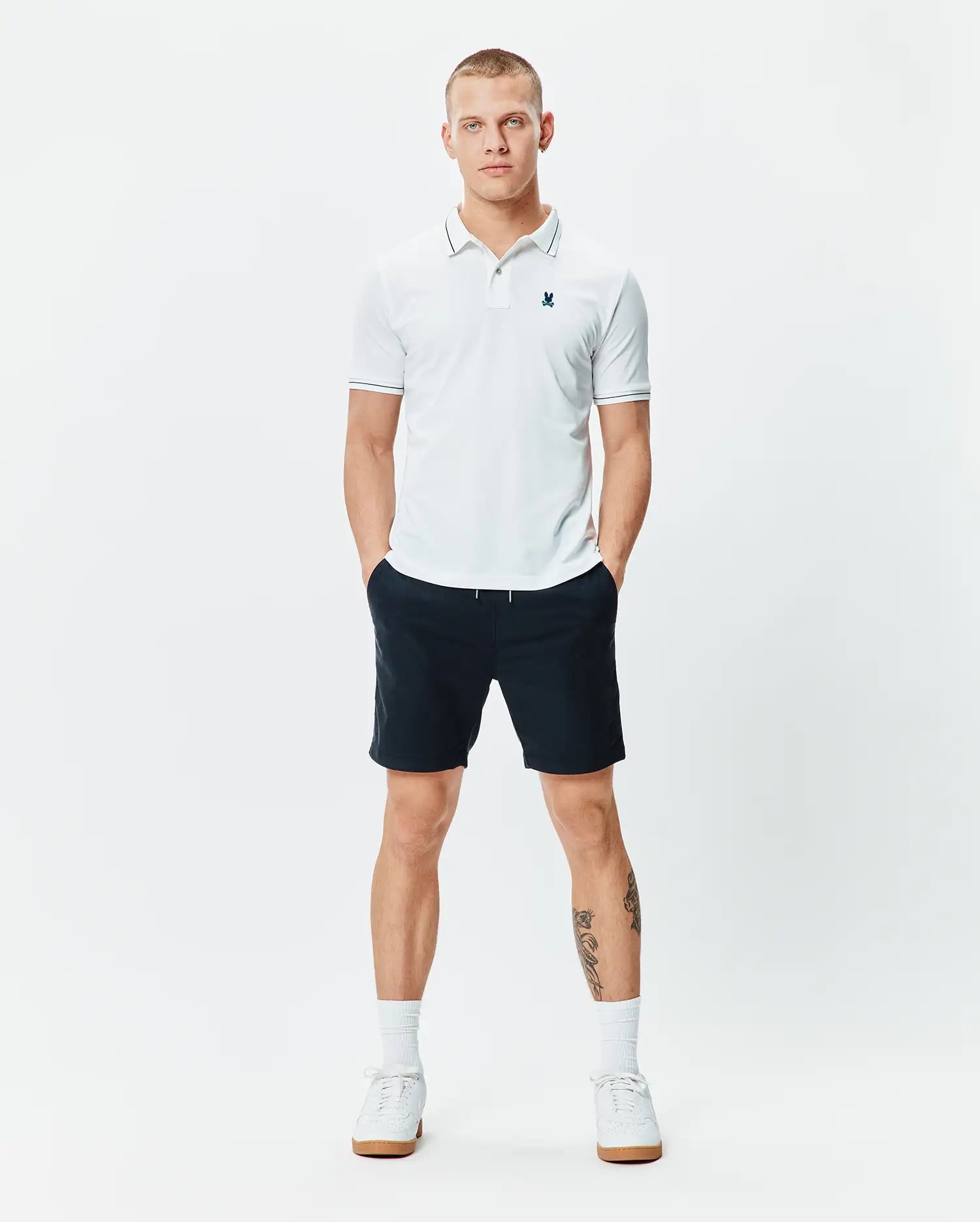 Mens Edwin Sport Polo, a classic fit white pique polo with navy tipping on the collar and cuffs, featuring a small embroidered teal Psycho Bunny skull logo on the left chest.