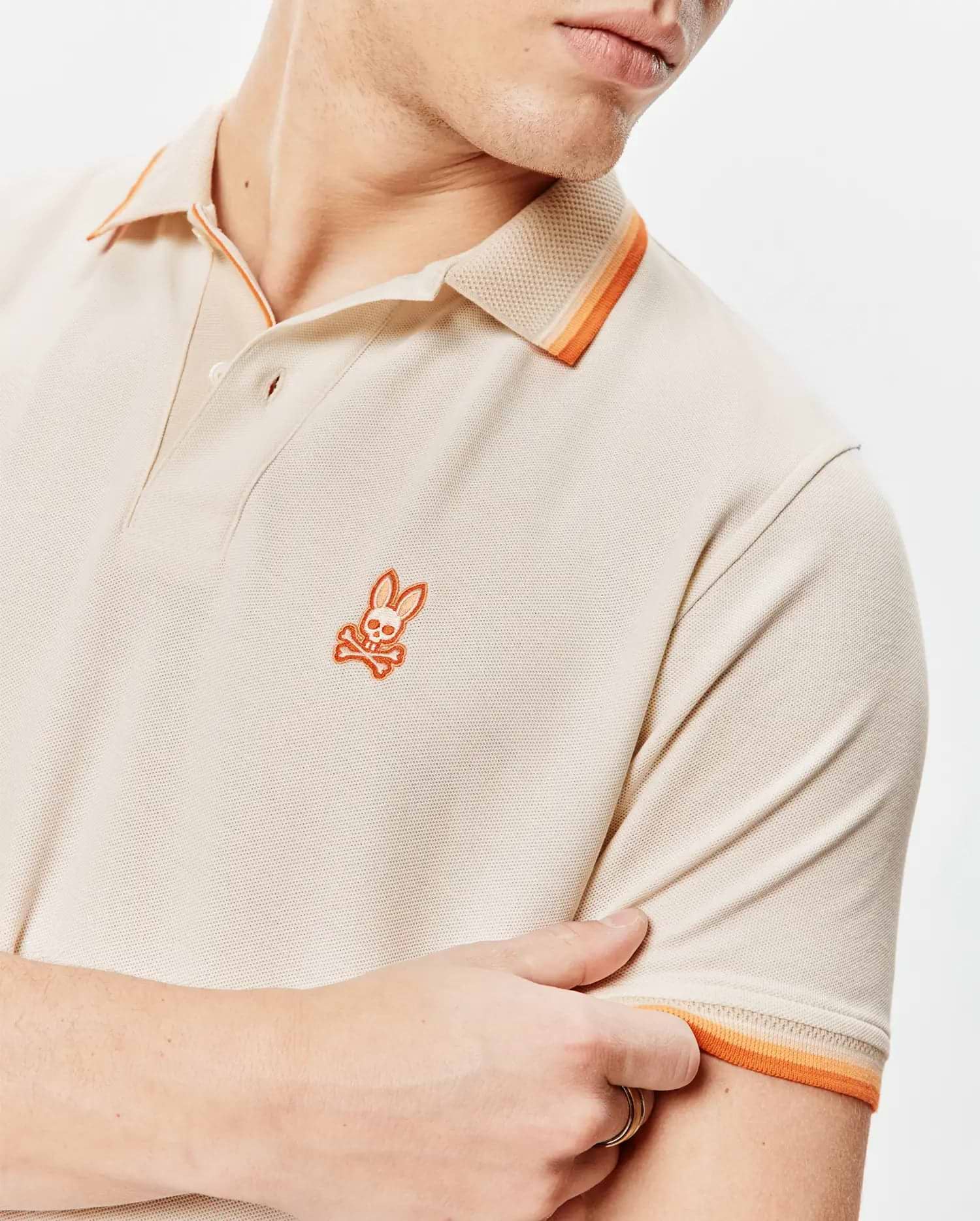 Mens Holden Pique Polo, a classic fit beige pique polo shirt featuring a small embroidered orange Psycho Bunny skull logo on the left chest and striped orange collar and sleeve cuffs. (2)