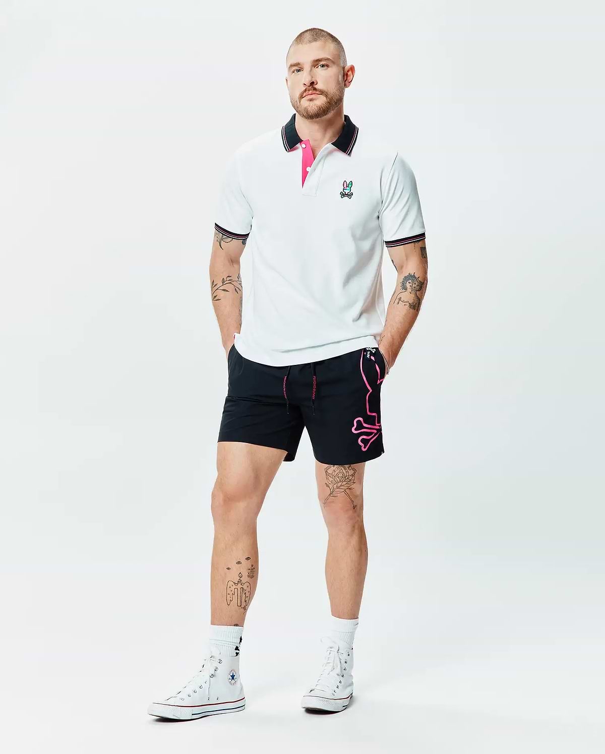 White Mens Nate Pique Polo with black collar, pink under-collar, striped cuffs, and embroidered Psycho Bunny logo, paired with black shorts featuring a large pink Psycho Bunny skull graphic.