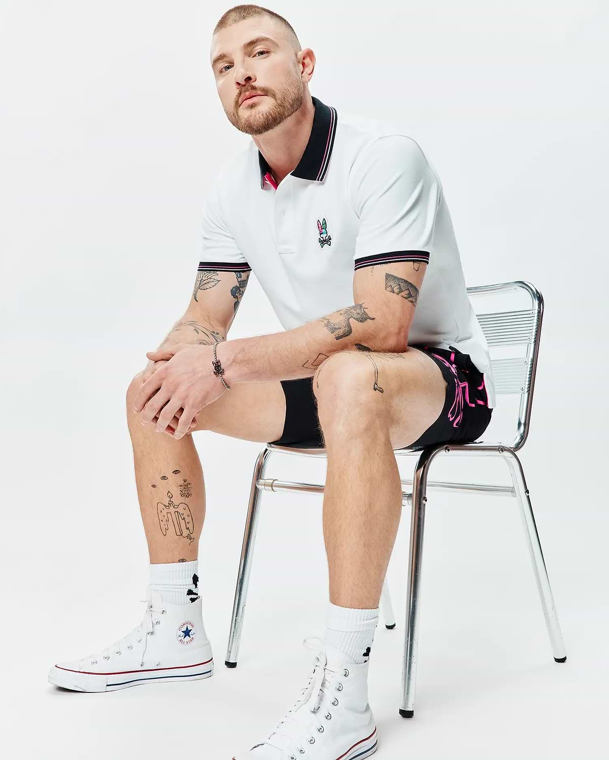 MENS NATE PIQUE POLO, a white classic pique polo with black and pink striped collar/cuffs, featuring a colorful embroidered Psycho Bunny logo. Black shorts with a vibrant pink Psycho Bunny side graphic.