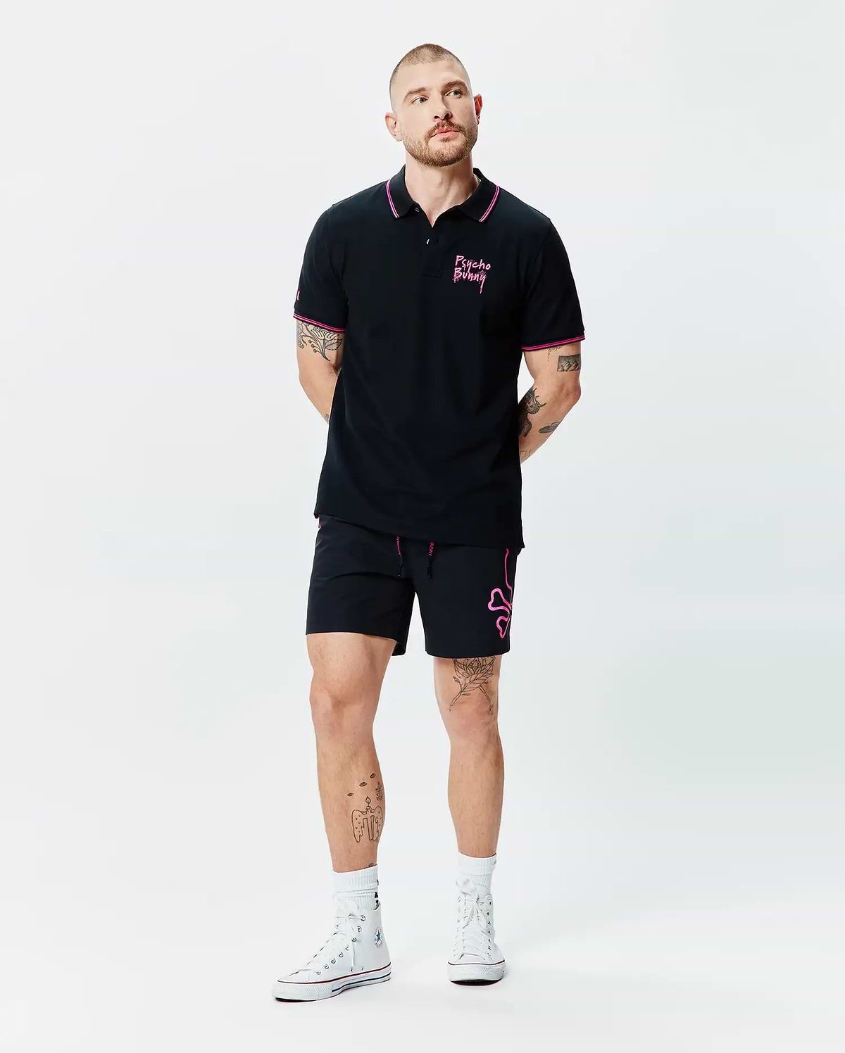 Black Mens Duncan Pique Polo with pink collar and cuff tipping, featuring a pink Psycho Bunny script logo on the left chest. Paired with black shorts showcasing a large pink bunny skull outline and pink drawstrings.