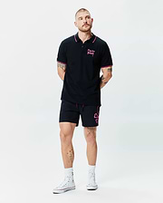 Black Mens Duncan Pique Polo with pink collar and cuff tipping, featuring a pink Psycho Bunny script logo on the left chest. Paired with black shorts showcasing a large pink bunny skull outline and pink drawstrings.