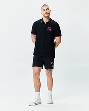 Black Mens Duncan Pique Polo with pink collar and cuff tipping, featuring a pink Psycho Bunny script logo on the left chest. Paired with black shorts showcasing a large pink bunny skull outline and pink drawstrings.