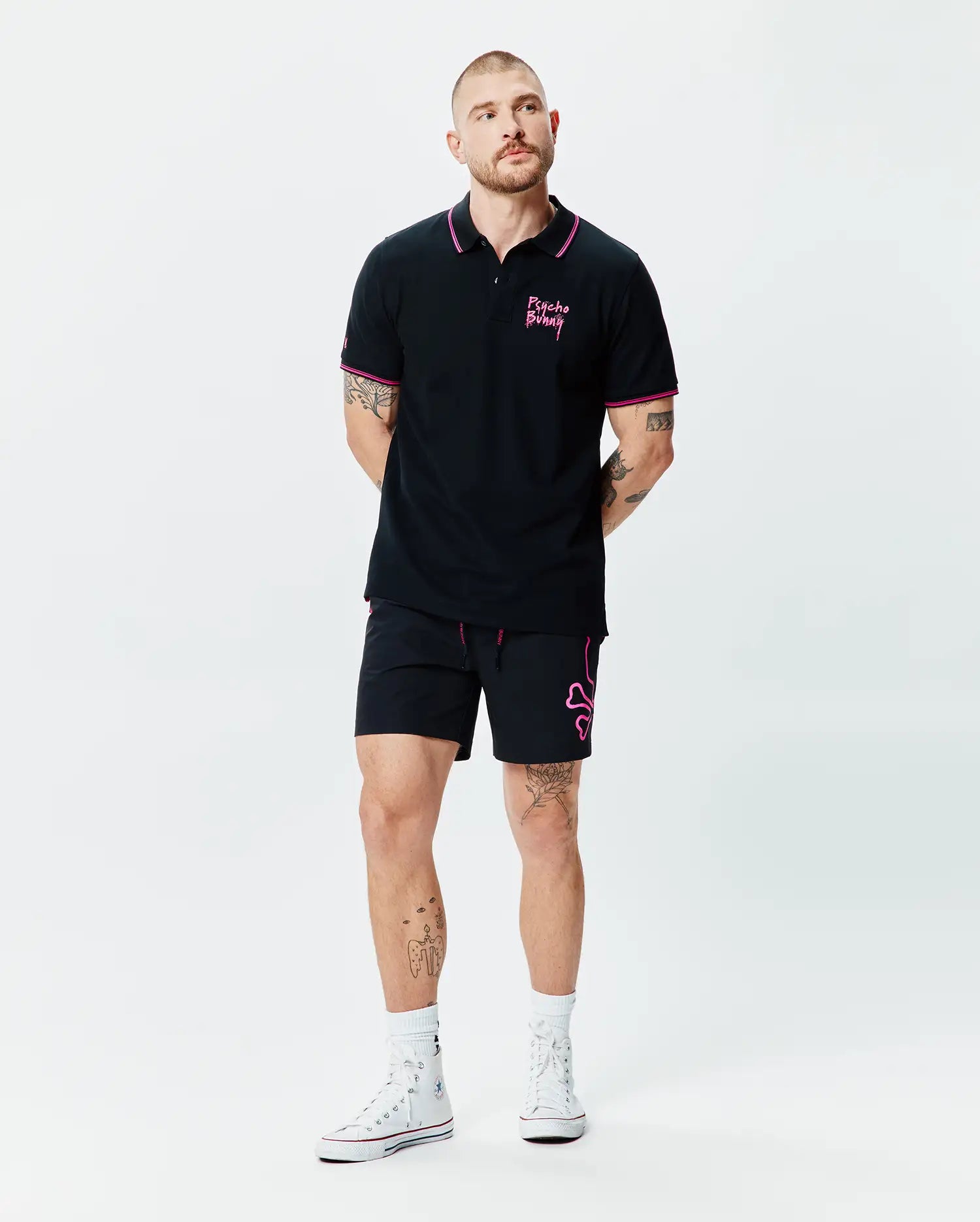 Black Mens Duncan Pique Polo with pink collar and cuff tipping, featuring a pink Psycho Bunny script logo on the left chest. Paired with black shorts showcasing a large pink bunny skull outline and pink drawstrings.