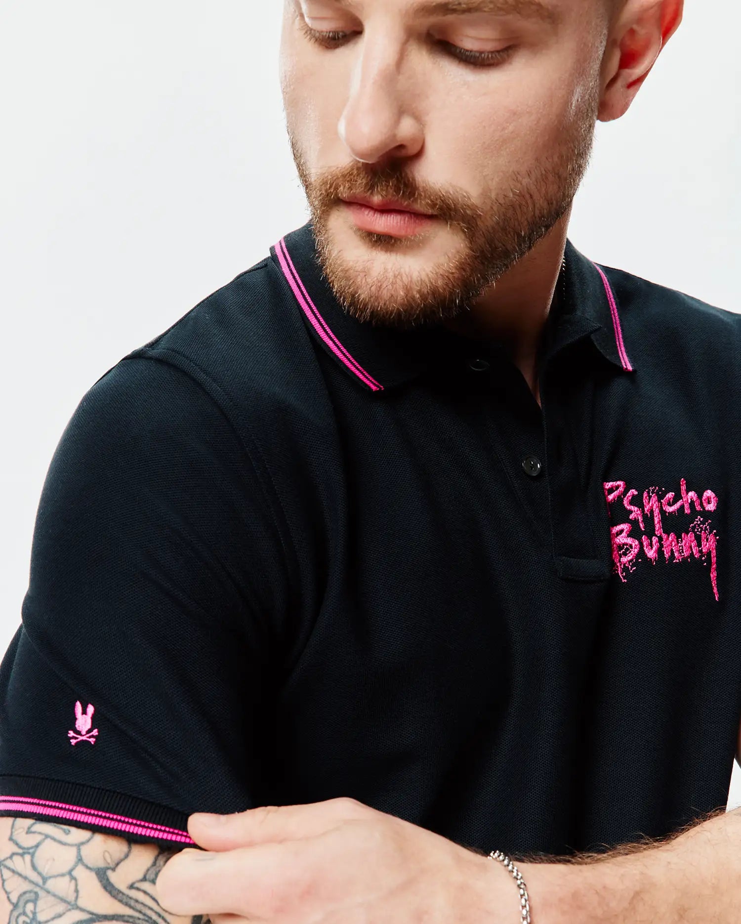 Mens Duncan Pique Polo, black classic fit pique polo with hot pink tipping on collar and cuffs, featuring a small embroidered pink bunny skull on the left sleeve and a large distressed pink Psycho Bunny text logo on the right chest.
