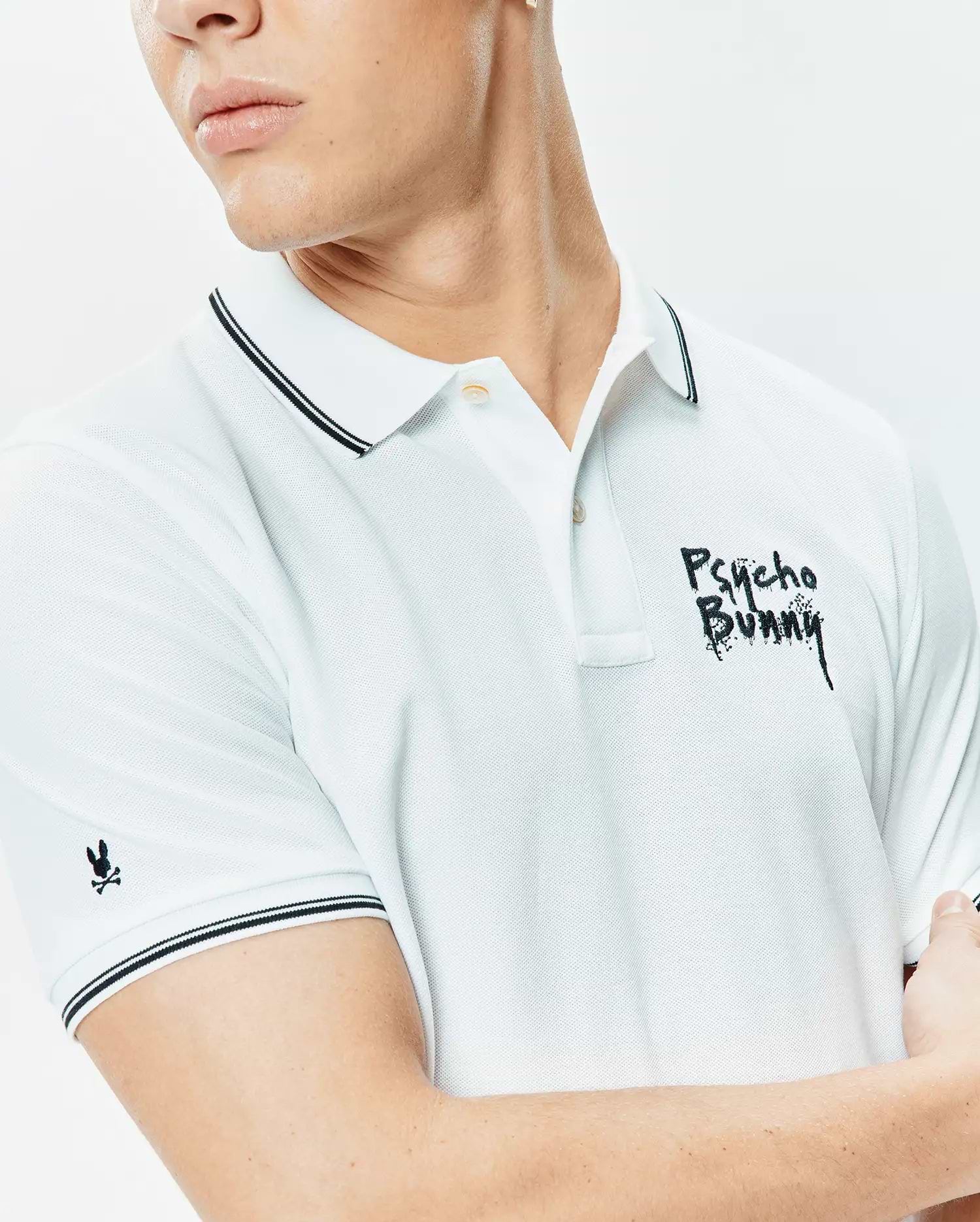 Mens Duncan Pique Polo, a classic fit white pique polo shirt with black double-tipped collar and cuffs, featuring a large embroidered black distressed Psycho Bunny wordmark on the chest and a small black bunny skull logo on the sleeve.