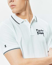 Mens Duncan Pique Polo, a classic fit white pique polo shirt with black double-tipped collar and cuffs, featuring a large embroidered black distressed Psycho Bunny wordmark on the chest and a small black bunny skull logo on the sleeve.