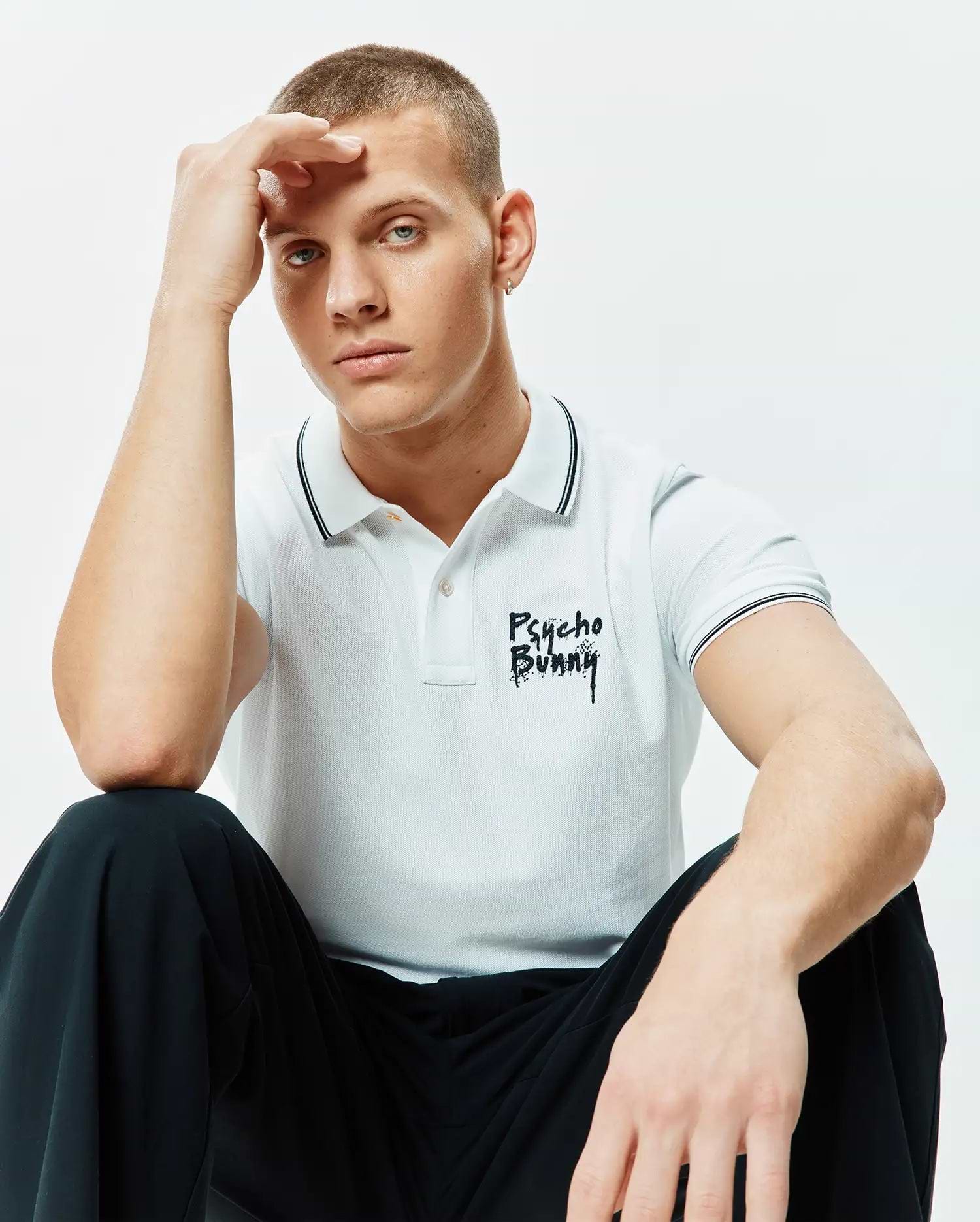 Mens Duncan Pique Polo, white classic fit pique knit polo with black double-tipped collar and cuffs, featuring a distressed black embroidered Psycho Bunny text logo on the left chest.