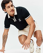 Black Mens Ridley Pique Polo with cream ribbed collar, placket, and striped cuffs, featuring an embroidered white Psycho Bunny skull logo on the chest, paired with cream waffle knit shorts. (3)
