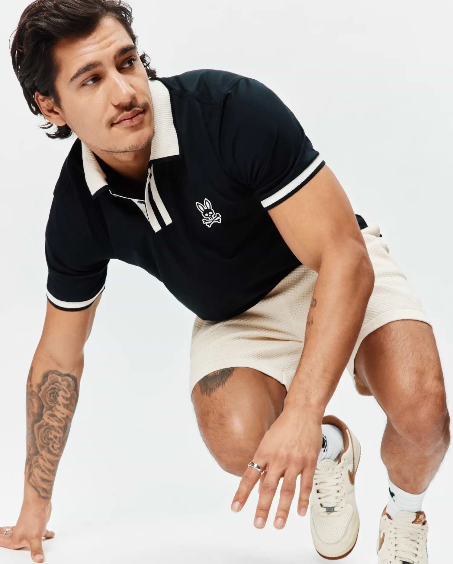 Black Mens Ridley Pique Polo with cream ribbed collar, placket, and striped cuffs, featuring an embroidered white Psycho Bunny skull logo on the chest, paired with cream waffle knit shorts. (3)