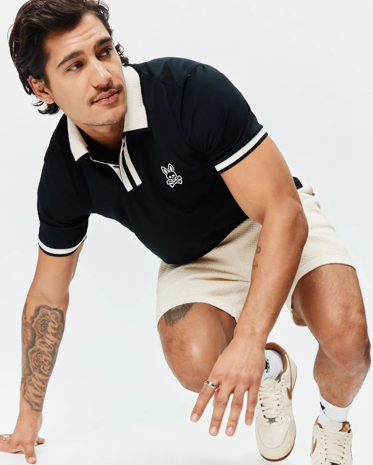Black Mens Ridley Pique Polo with cream ribbed collar, placket, and striped cuffs, featuring an embroidered white Psycho Bunny skull logo on the chest, paired with cream waffle knit shorts. (3)