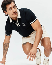 Black Mens Ridley Pique Polo with cream ribbed collar, placket, and striped cuffs, featuring an embroidered white Psycho Bunny skull logo on the chest, paired with cream waffle knit shorts. (3)