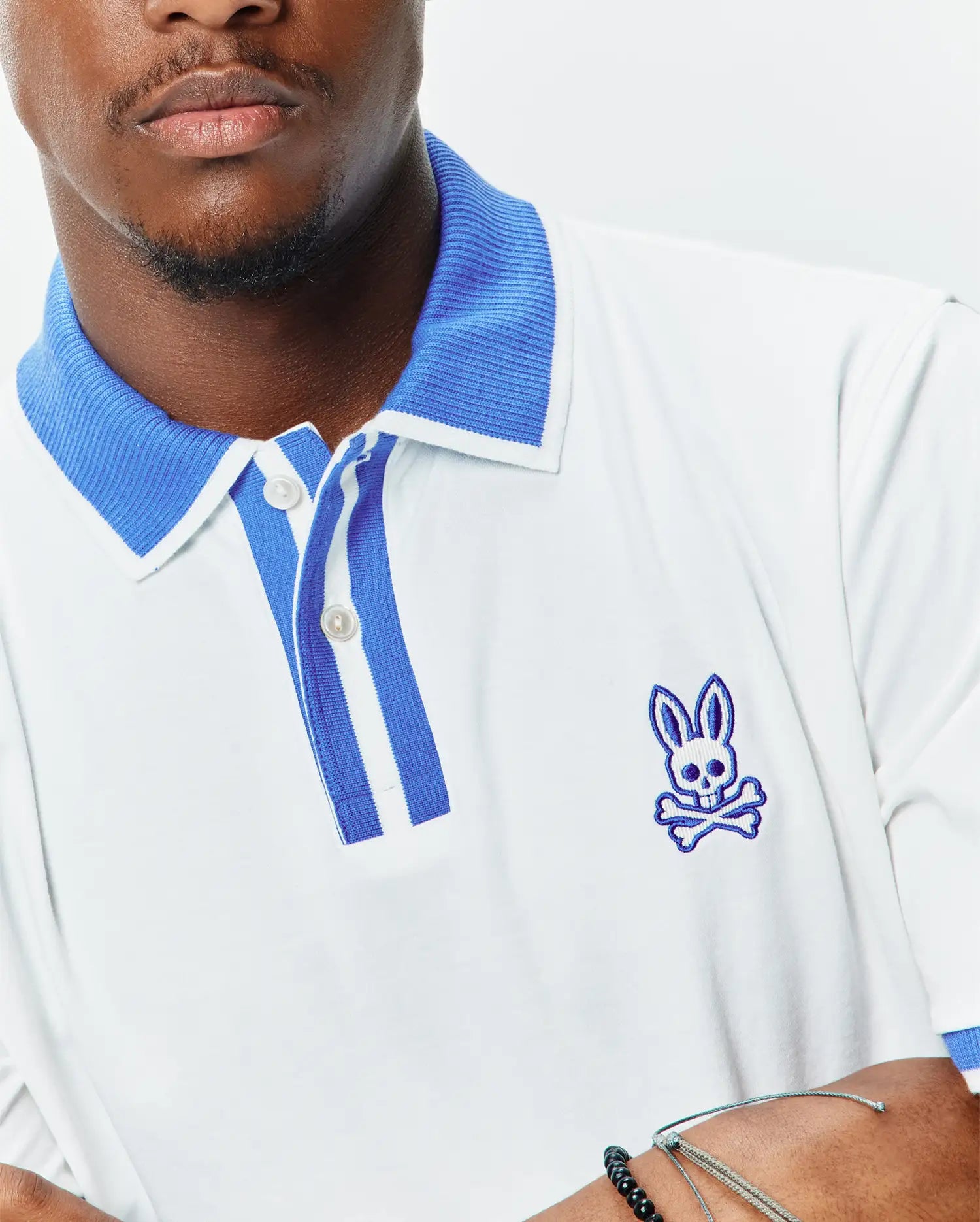 Mens Ridley Pique Polo in white pique, featuring a classic fit, ribbed blue collar, striped blue and white placket, and an embroidered blue Psycho Bunny skull logo on the left chest. (2)