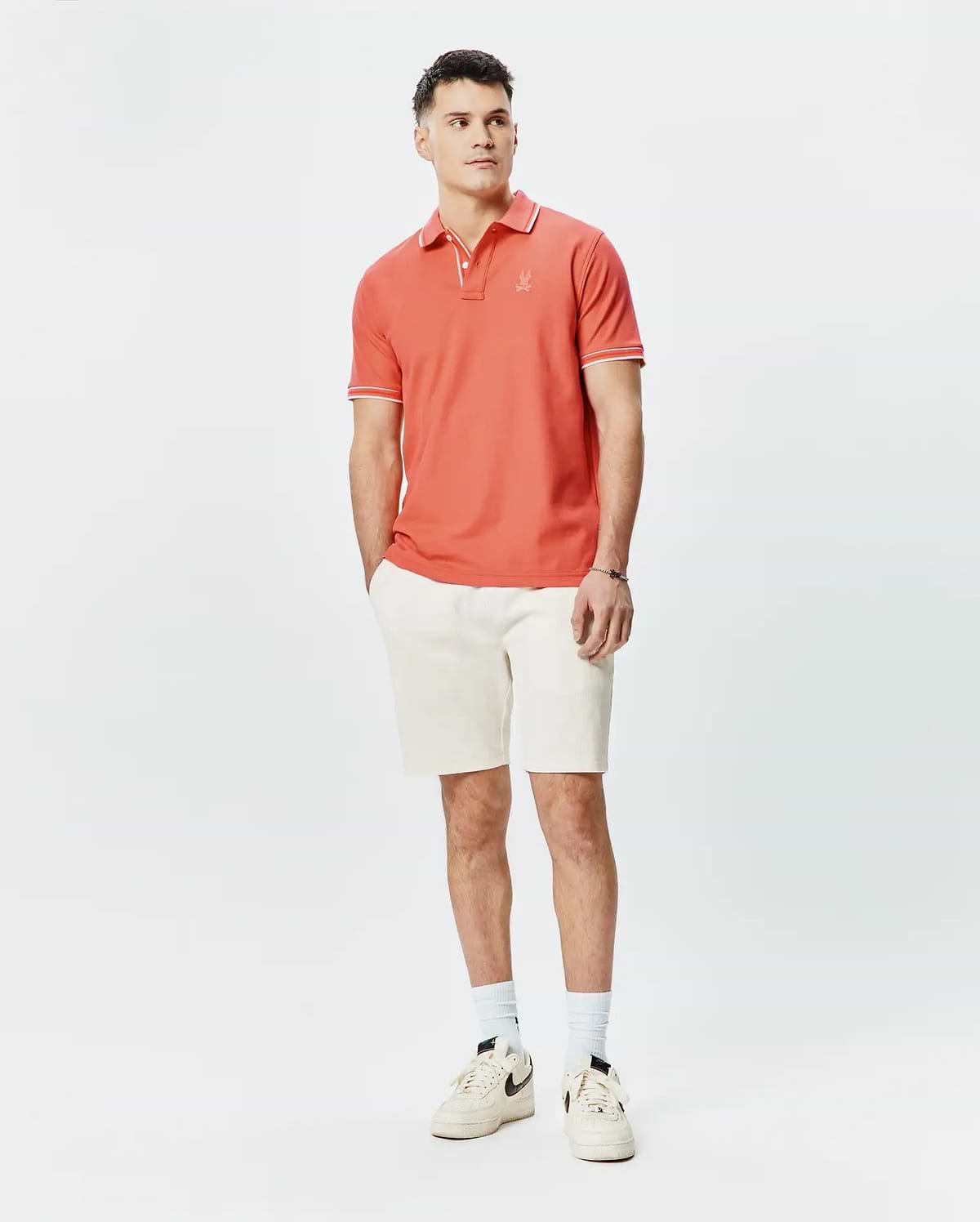 Mens Brad Pique Polo in vibrant coral, classic fit, with white tipping on collar and short sleeves, featuring a small embroidered Psycho Bunny skull logo on the left chest, paired with cream textured shorts. (4)