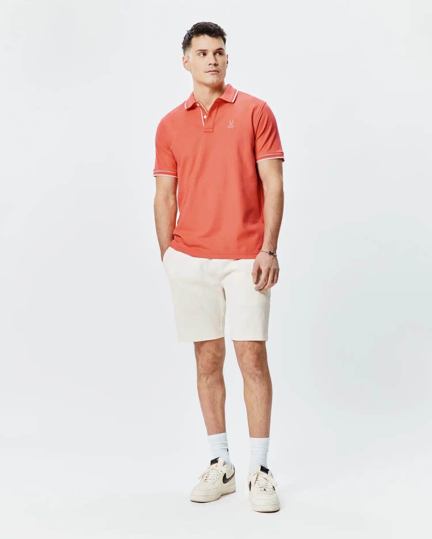 Mens Brad Pique Polo in vibrant coral, classic fit, with white tipping on collar and short sleeves, featuring a small embroidered Psycho Bunny skull logo on the left chest, paired with cream textured shorts. (4)