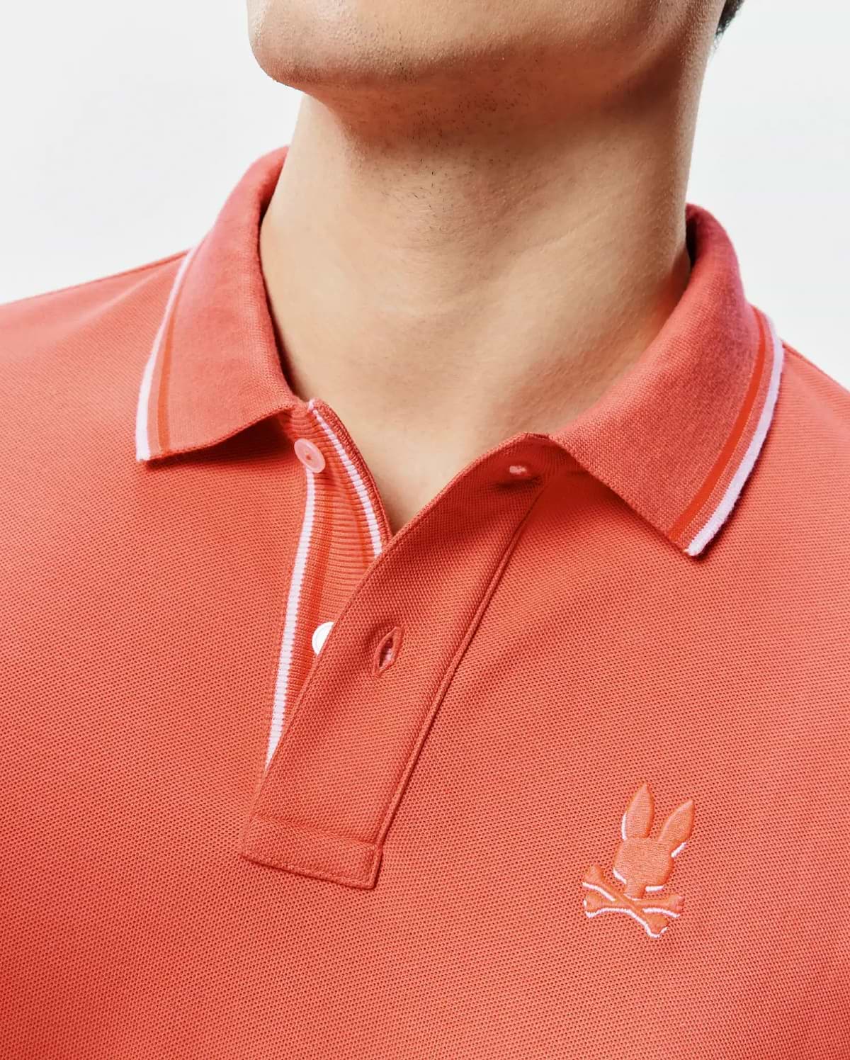 Mens Brad Pique Polo in vibrant coral, featuring a classic fit, textured pique knit, a ribbed collar with white and orange stripes, and a small embroidered Psycho Bunny skull logo on the left chest. (2)