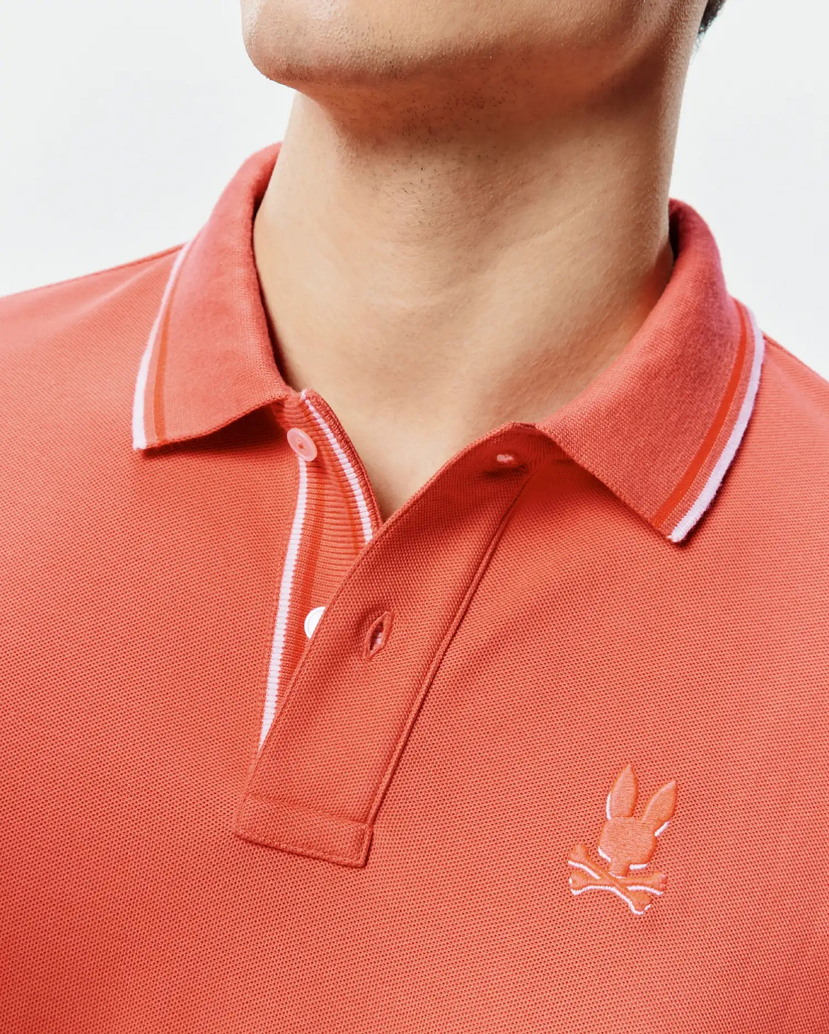 Mens Brad Pique Polo in vibrant coral, featuring a classic fit, textured pique knit, a ribbed collar with white and orange stripes, and a small embroidered Psycho Bunny skull logo on the left chest. (2)