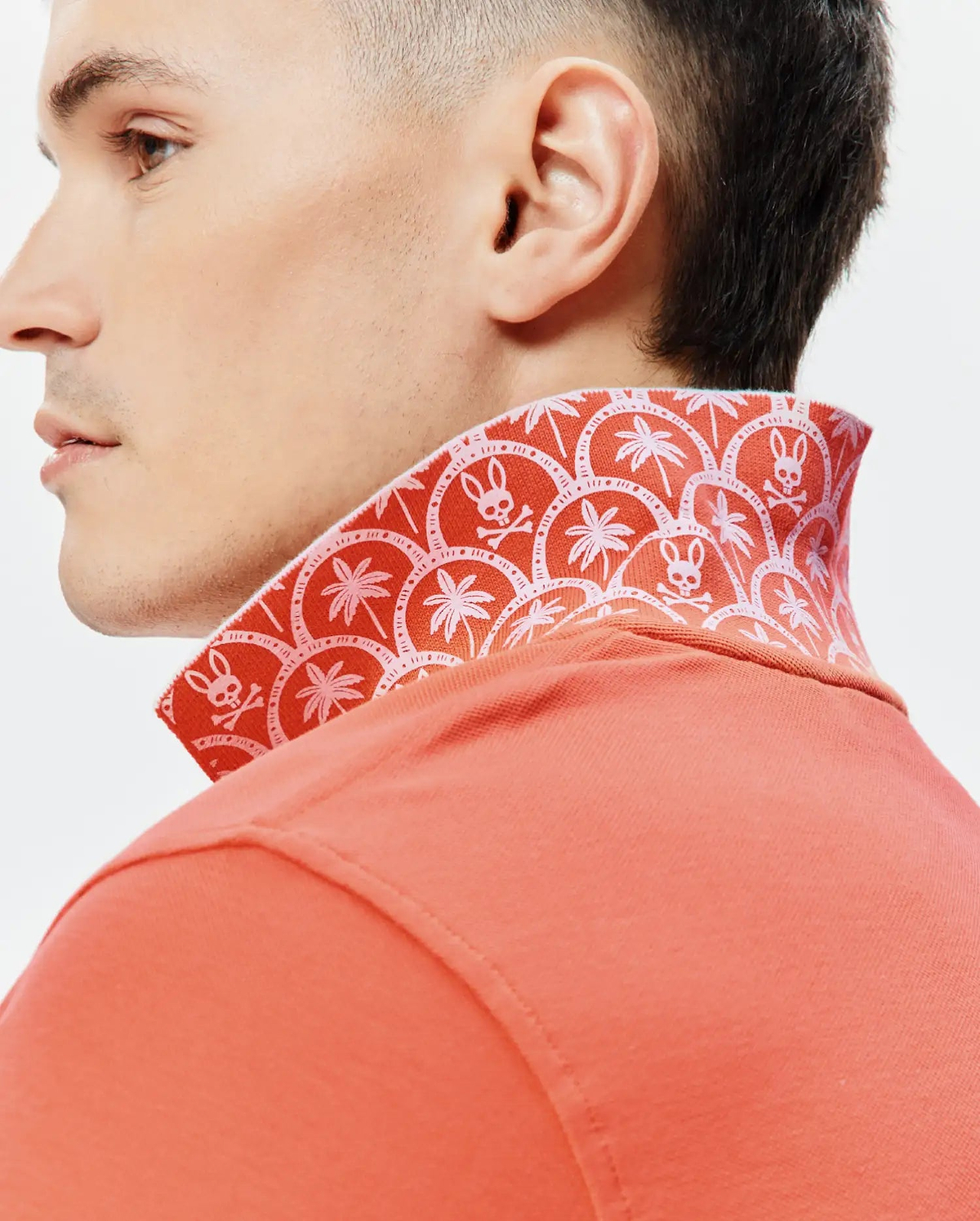 Mens Brad Pique Polo in coral pique, featuring a vibrant red-orange patterned collar with white Psycho Bunny skull logos and palm trees, embodying refined streetwear. (3)