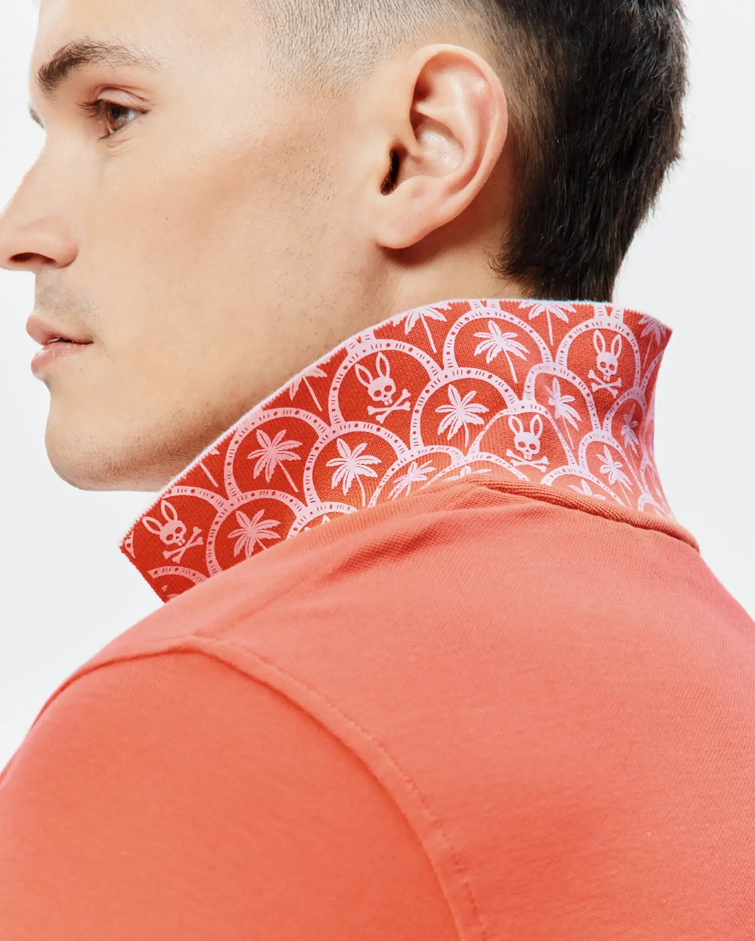 Mens Brad Pique Polo in coral pique, featuring a vibrant red-orange patterned collar with white Psycho Bunny skull logos and palm trees, embodying refined streetwear. (3)