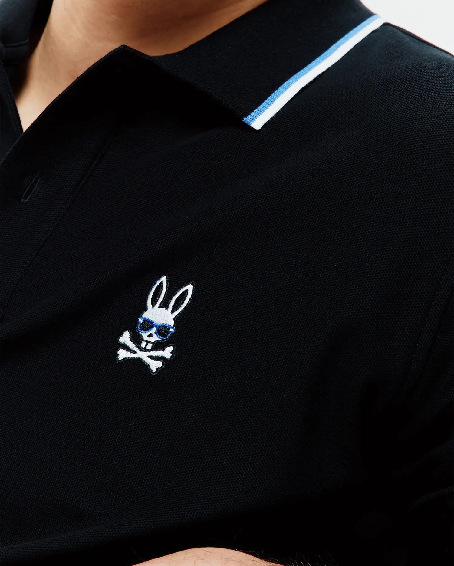 Mens Sunny Pique Polo, black classic fit pique polo with a ribbed collar featuring white and blue stripes, and a small embroidered Psycho Bunny skull logo with blue sunglasses on the left chest. (2)