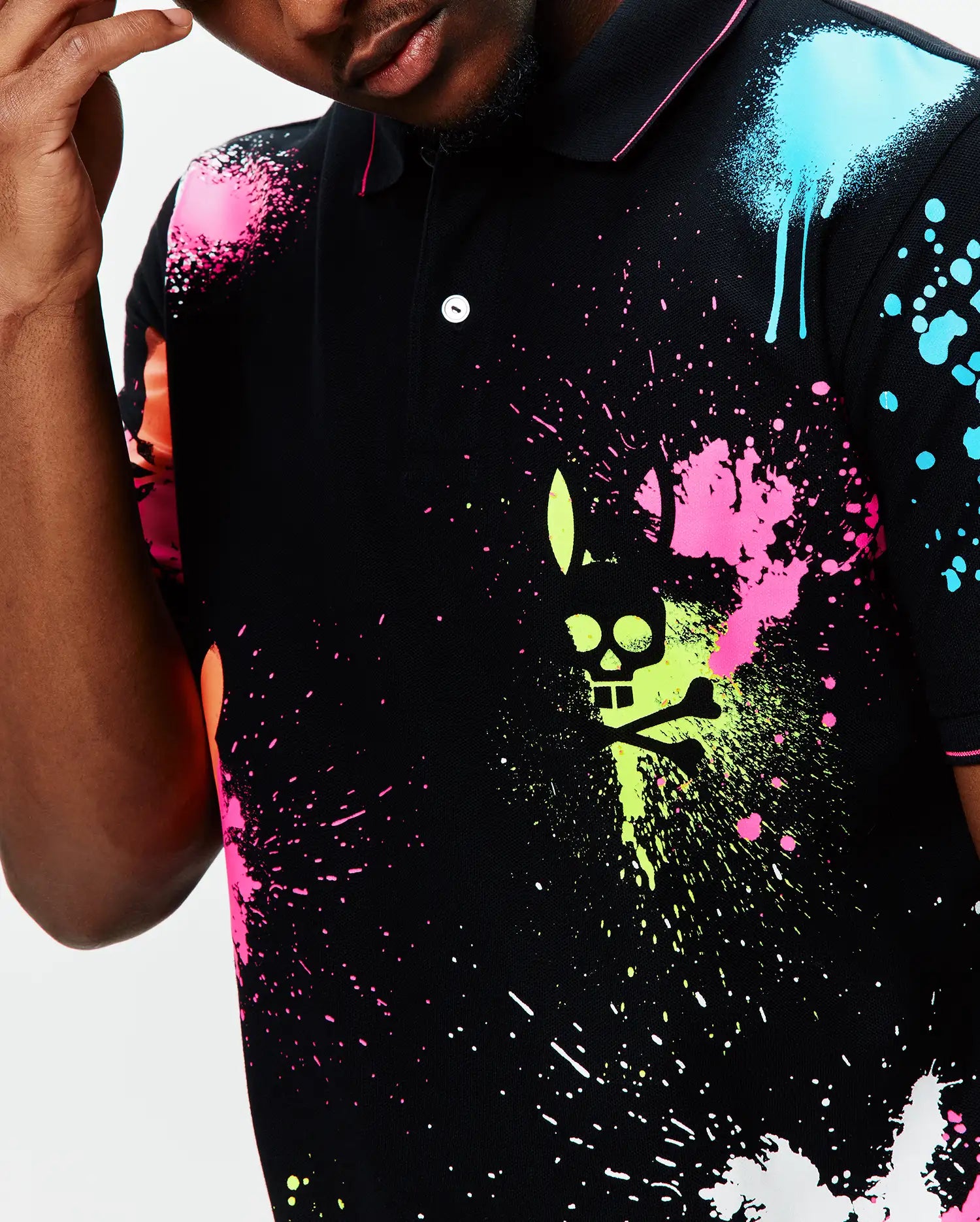 Mens Quintin Aop Pique Polo, a black pique polo with a vibrant all-over neon paint splatter print in pink, lime, blue, and orange, featuring a large central Psycho Bunny skull logo graphic.