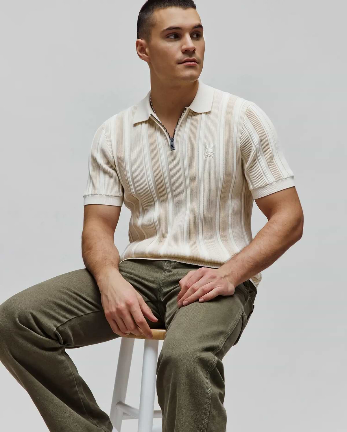 Mens Cliff Knit Zip Polo in cream and beige vertical stripes, featuring a ribbed knit texture, a quarter-zip closure with a Psycho Bunny logo pull, and a small embroidered cream bunny skull logo on the left chest for a slim fit.