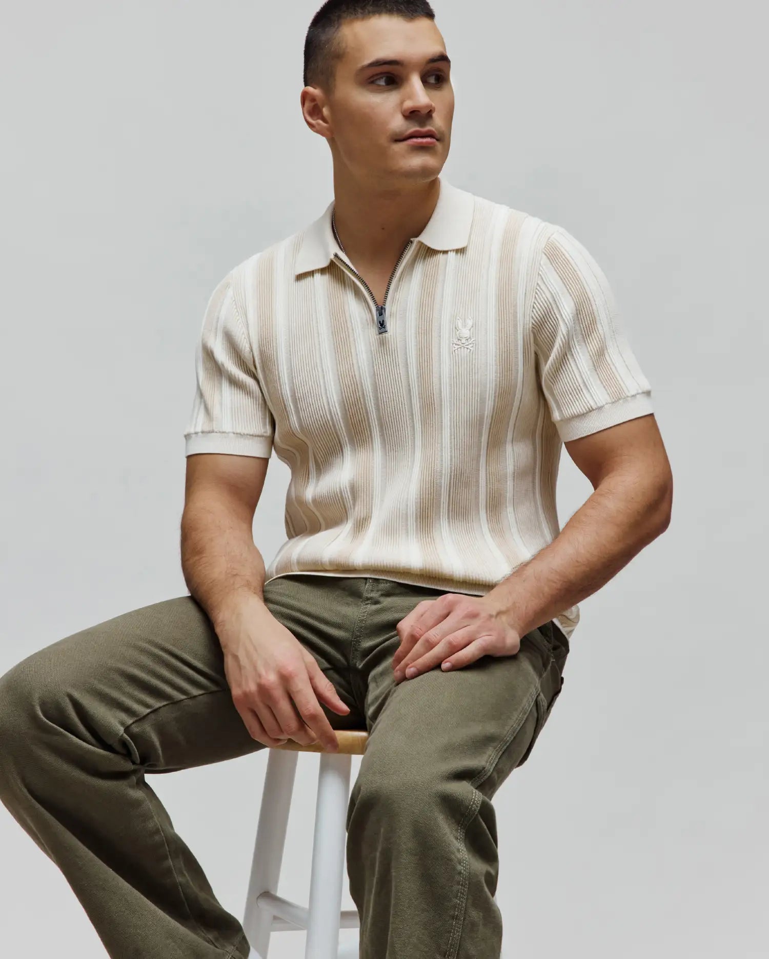 Mens Cliff Knit Zip Polo in cream and beige vertical stripes, featuring a ribbed knit texture, a quarter-zip closure with a Psycho Bunny logo pull, and a small embroidered cream bunny skull logo on the left chest for a slim fit.
