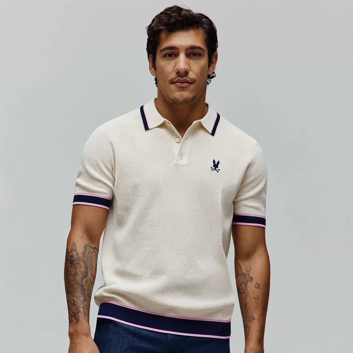 Mens Dax Knit Polo, cream textured knit with navy and pink striped collar, cuffs, and hem, featuring a small embroidered navy Psycho Bunny skull logo on the left chest. (2)