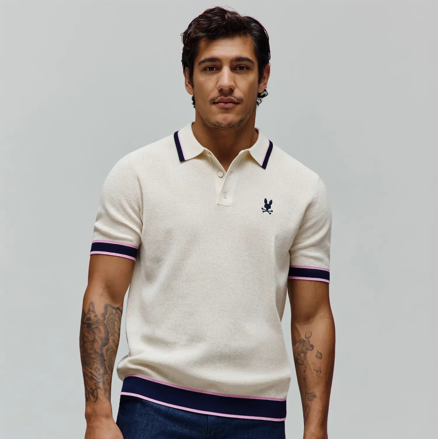 Mens Dax Knit Polo, cream textured knit with navy and pink striped collar, cuffs, and hem, featuring a small embroidered navy Psycho Bunny skull logo on the left chest. (2)
