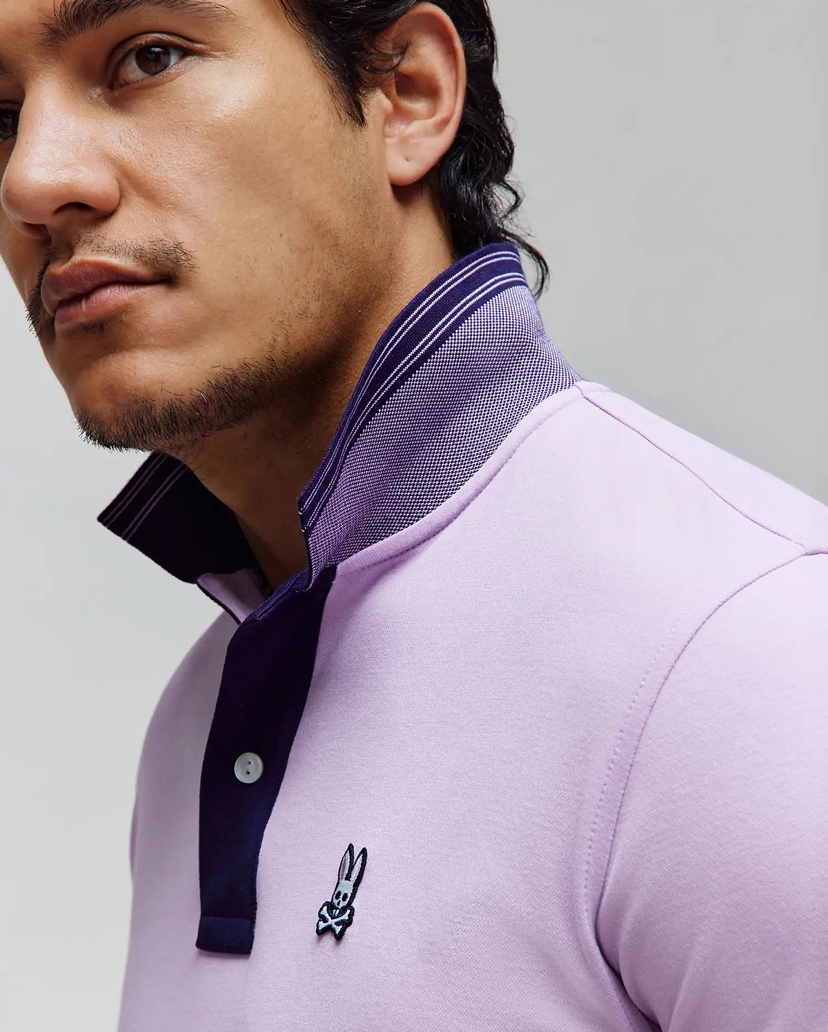 Mens Kenyon Pique Polo, a classic fit lavender pique polo with a dark purple two-button placket, striped collar, and small embroidered Psycho Bunny skull logo on the left chest.