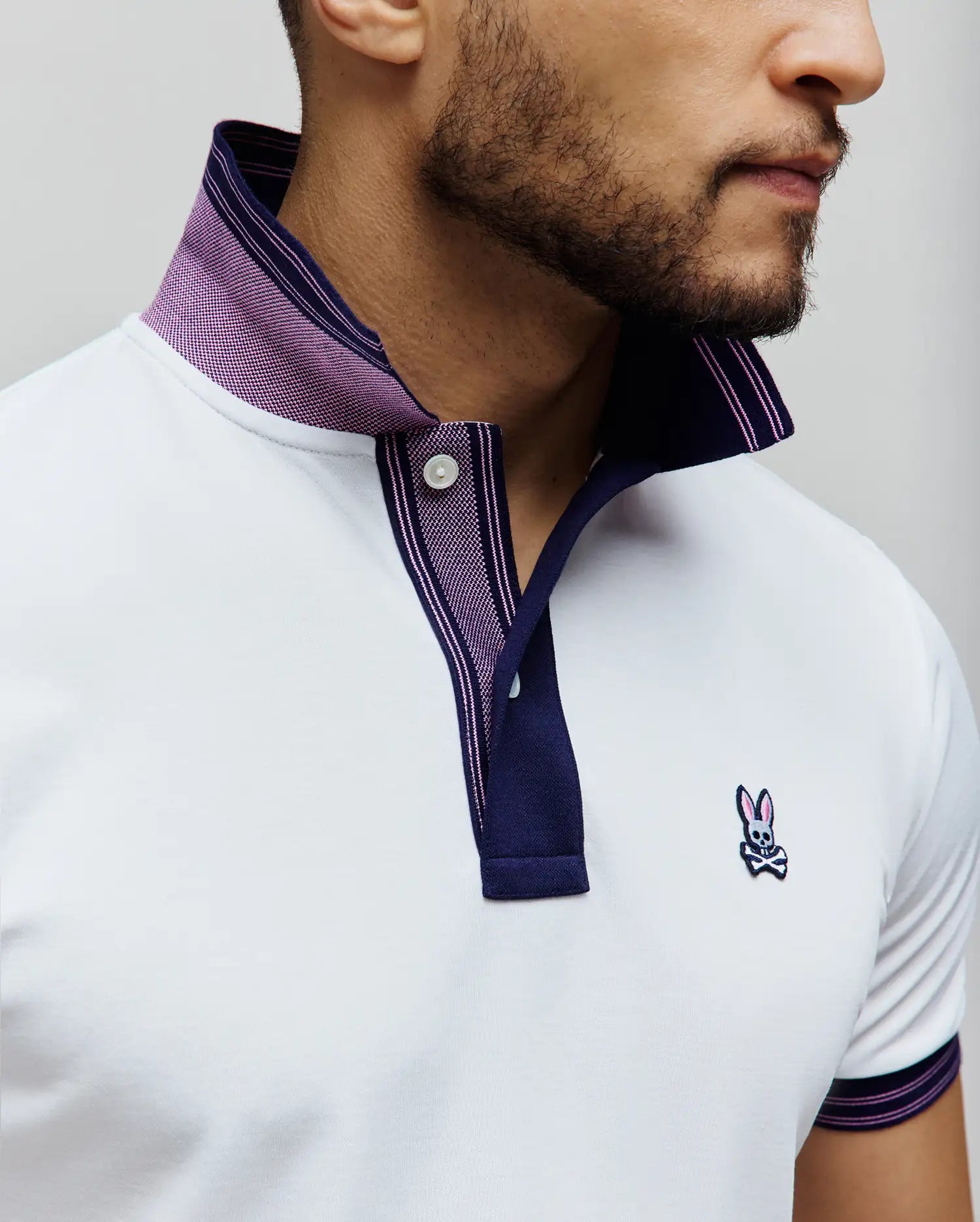 MENS KENYON PIQUE POLO, a white classic fit pique polo with navy and purple striped collar, placket, and sleeve cuffs, featuring a purple patterned inner collar and an embroidered Psycho Bunny skull logo on the left chest.