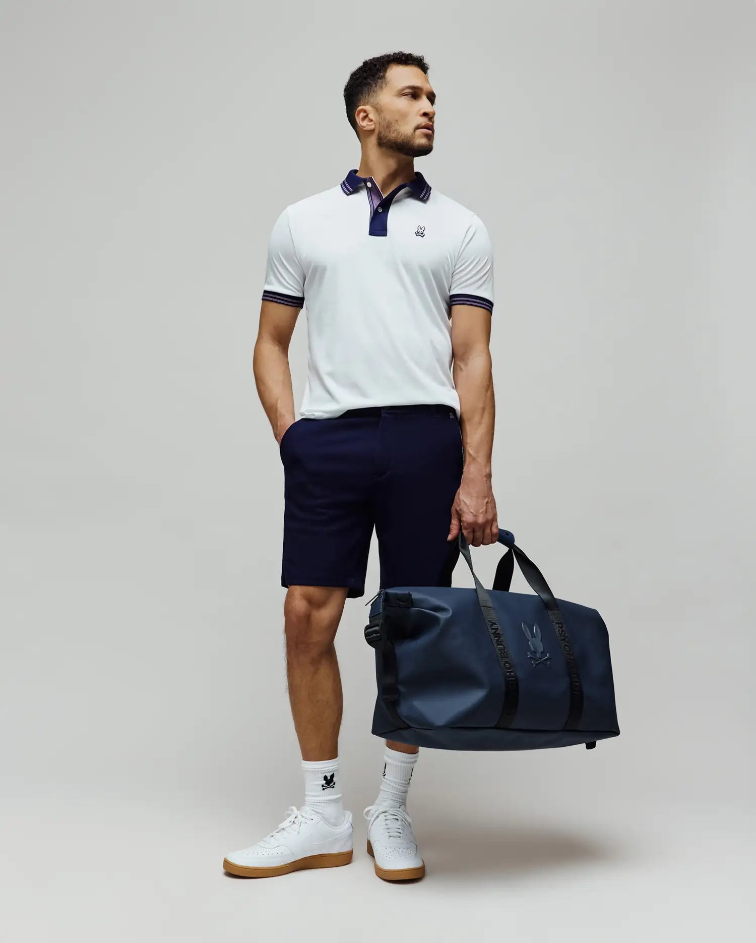 Mens Kenyon Pique Polo, white pique with navy collar, striped cuffs, and embroidered Psycho Bunny logo. Styled with navy shorts, white logo socks, white sneakers, and a navy Psycho Bunny duffel bag.