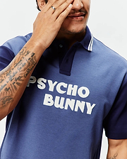 Mens Kelly Oversized Polo, a color-block jersey polo in faded blue with navy sleeves and collar, featuring white collar stripes and a large textured white appliqué PSYCHO BUNNY logo on the chest. (3)