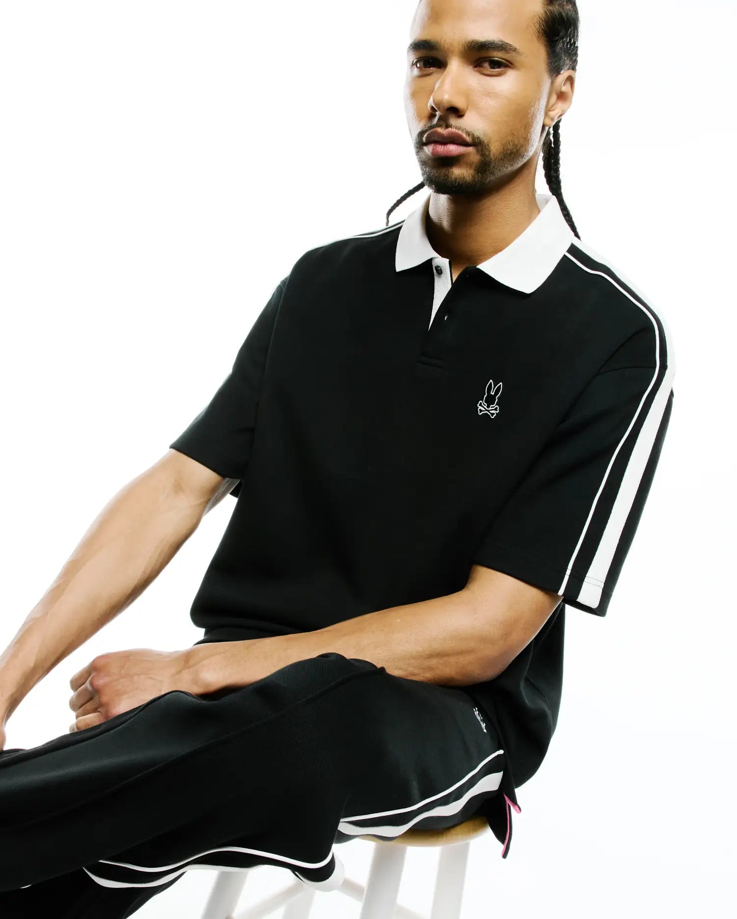 Mens Ceano Relaxed Fit Heavy Pique Polo in black with a white collar, white double stripes on sleeves, small embroidered white bunny skull logo on left chest, and side vents with a pink accent.
