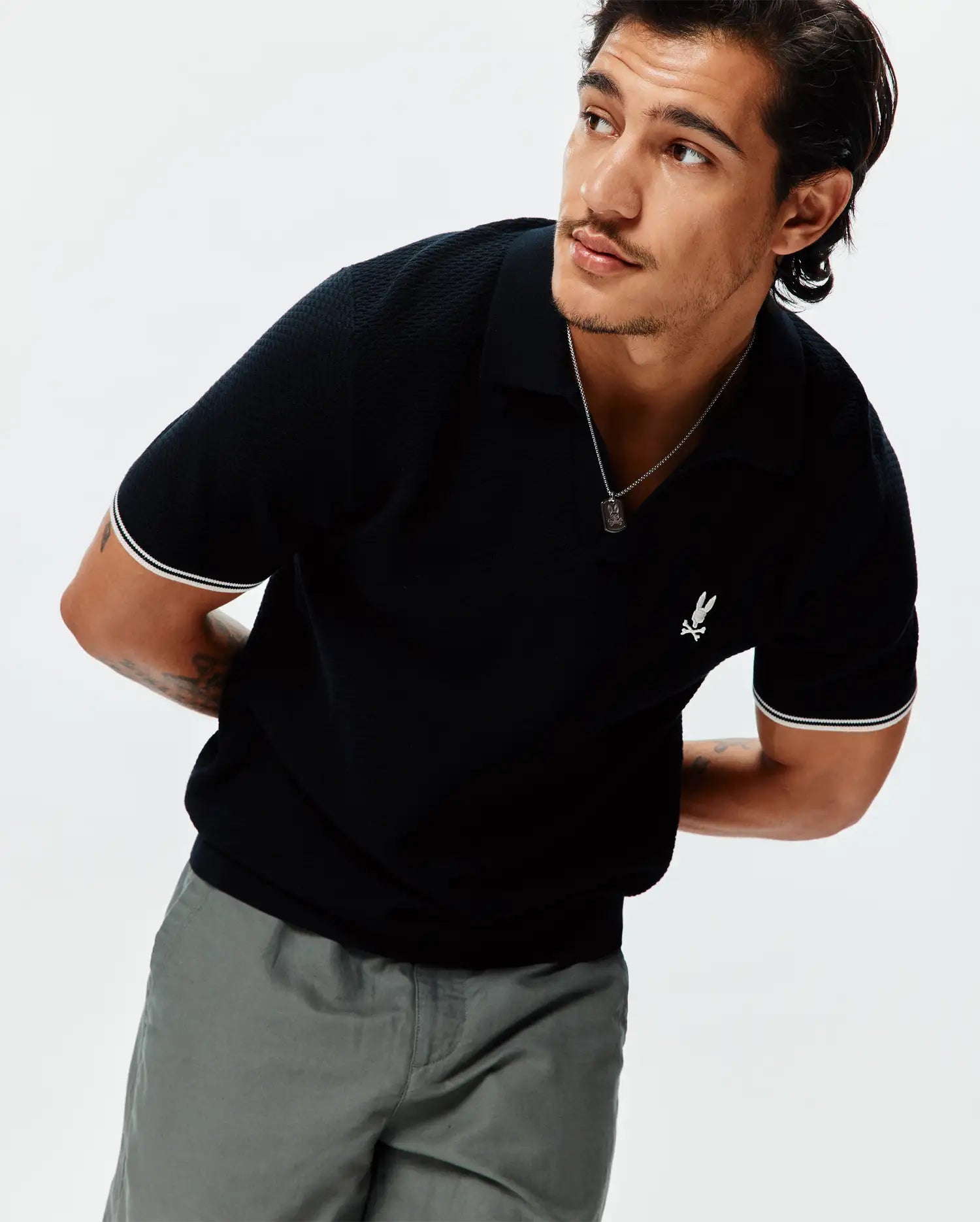 Mens Selby Knit Polo, black textured knit with white double-stripe ribbed cuffs and a small white embroidered Psycho Bunny skull logo on the left chest, classic fit. (1)